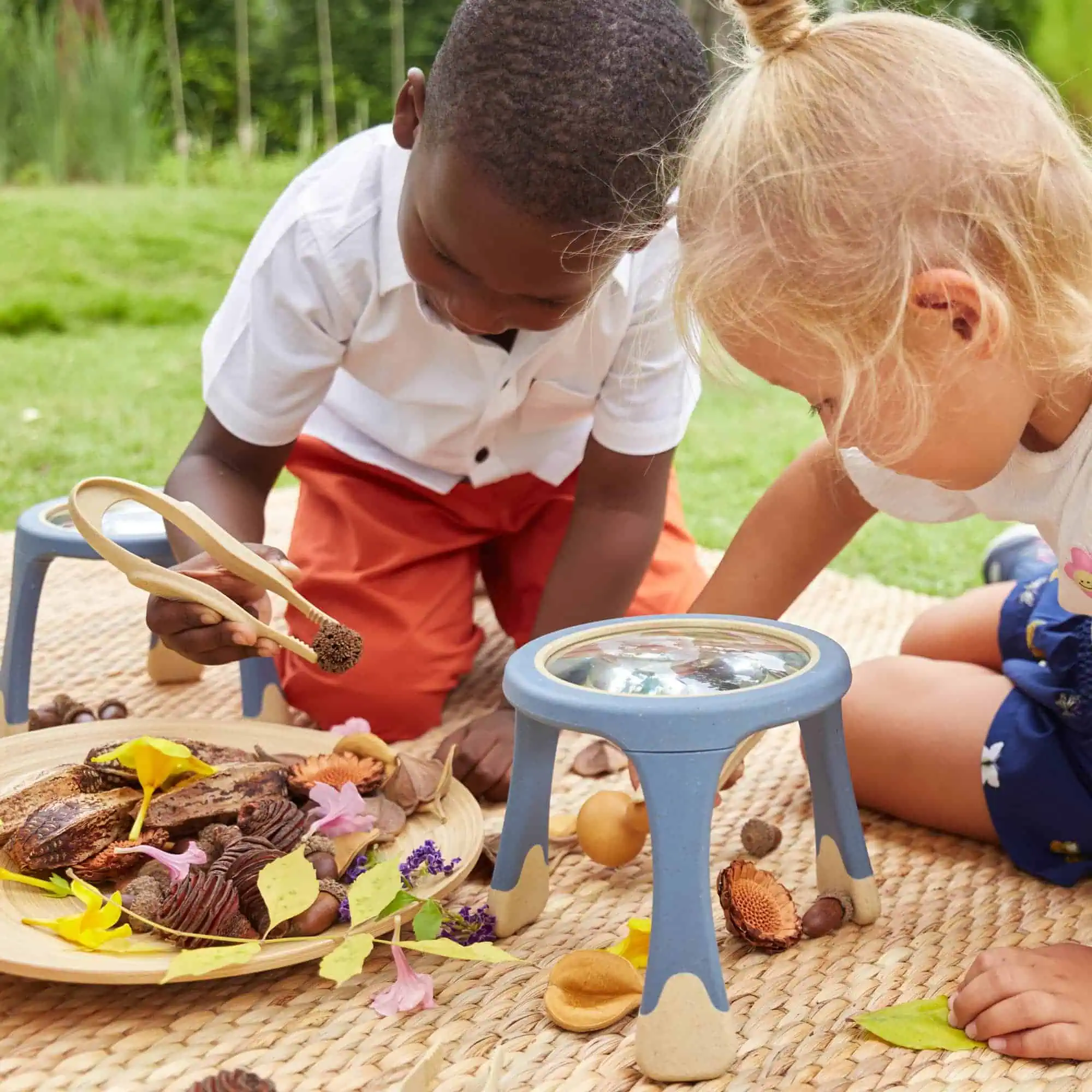 Im Bild sehen zwei Kinder, die draußen auf einer Decke mit Büchern und Naturmaterialien verschiedene Experimente durchführen und die Natur neugierig erkunden.